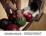 Person selecting bowling ball from ball return system at bowling alley, preparing for next turn at lane, visible in casual clothing, showcasing hands and equipment