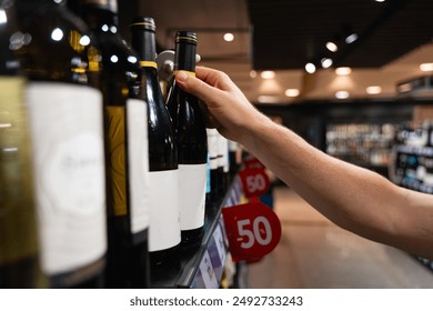 A person selects a bottle of wine from a supermarket shelf, with a focus on their hand and the wine bottles. male consumer concept  - Powered by Shutterstock