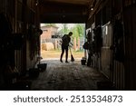 Person in silhouette cleaning barn floor with a broom, surrounded by equipment and horse stalls, sunlight streaming through open barn door leading outside