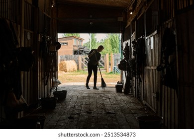 Person in silhouette cleaning barn floor with a broom, surrounded by equipment and horse stalls, sunlight streaming through open barn door leading outside - Powered by Shutterstock