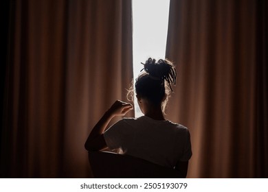 Person silhouetted sitting against light while peering through curtains. - Powered by Shutterstock