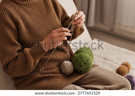 A person sits on a couch, using knitting needles to create a piece with different colors of yarn nearby. The room has natural light coming in from a window.