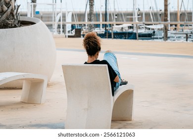 A person sitting back on a contemporary concrete bench, enjoying the view of the marina with palm trees and sailboats in the background. - Powered by Shutterstock