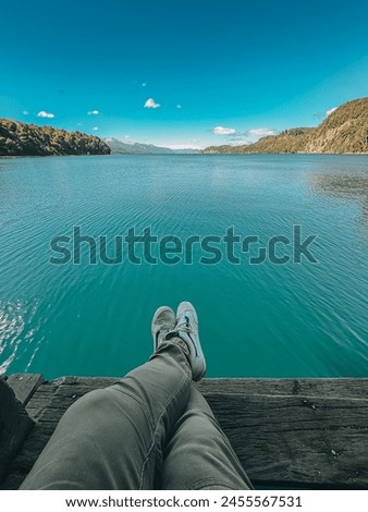 A person is sitting on a dock by a lake, looking out at the water. The scene is peaceful and serene, with the person's feet resting on the dock and the water reflecting the blue sky