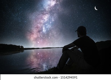 A Person Sitting On A Hill Looking At The Milky Way Galaxy Over A Lake