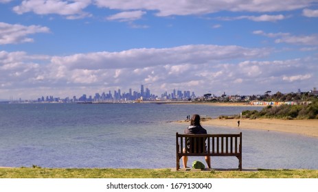 A person is sitting on the park bench of Brighton beach and looking at the Melbourne skyline.