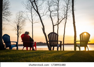 A Person Sitting In One Of Four Adirondack Chairs, Overlooking The Lake At Sunset On A Clear Day. Silhouette Of Chairs And Trees.