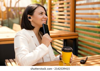 A Person Is Sitting At A Table Outside In A Cafe With A Phone In His Hand And A Thoughtful Look. A Woman Drinks Coffee, Listen To Music And Enjoy Life In Peace. Wireless Mini Headphones