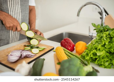 A person skillfully chops cucumbers and onions on a cutting board, surrounded by vibrant vegetables including lettuce, peppers, and corn in a sunny kitchen. - Powered by Shutterstock