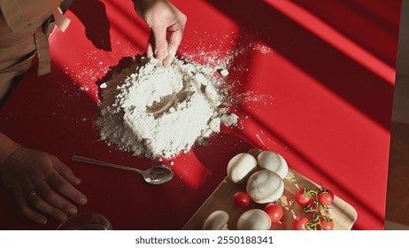 A person skillfully prepares pizza dough on a vibrant red surface, surrounded by fresh ingredients including mushrooms and cherry tomatoes, creating a lively culinary scene. - Powered by Shutterstock