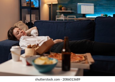 Person sleeping on sofa after drinking beer and eating home delivery pizza in front of television in living room. Tired woman falling asleep couch after large fast food takeout meal. - Shutterstock ID 2086013689