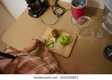 A person slices fresh fruits and vegetables on a wooden board in a kitchen for a smoothie. The scene includes a blender, a kiwi, apple, and celery, highlighting healthy and organic ingredients. - Powered by Shutterstock