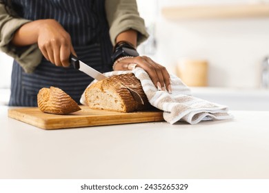 Person slices a loaf of bread on a wooden cutting board in a bright kitchen with copy space. Wearing a striped apron, the focus is on the hands and the bread, conveying a sense of home cooking. - Powered by Shutterstock