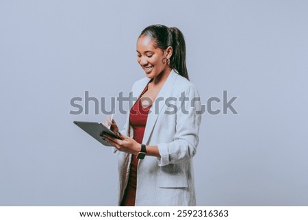 Person smiling while using a tablet, dressed in a light blazer and red top, standing against a plain background, engaged in digital work or note-taking. Business image.