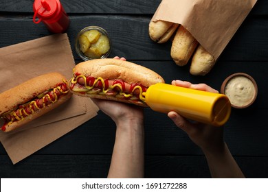 Person squeezes sauce on hot dog on wooden background, top view