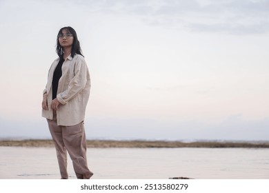 A person standing barefoot on a rocky shore covered with green seaweed, reflecting in shallow water during sunset. The background features a calm sea and a pastel sky. - Powered by Shutterstock