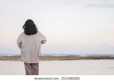 A person standing barefoot on a rocky shore covered with green seaweed, reflecting in shallow water during sunset. The background features a calm sea and a pastel sky. - Powered by Shutterstock