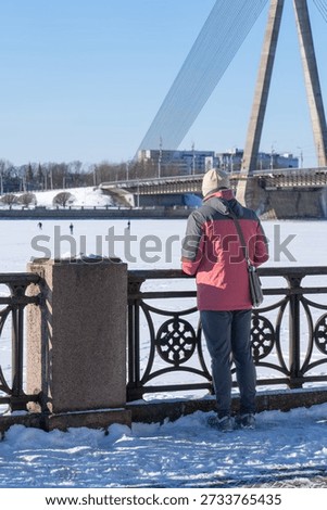Person standing by river railing overlooking frozen water and cable stayed bridge in winter city landscape, calm urban scene, 02.02.2026. Riga. Latvia