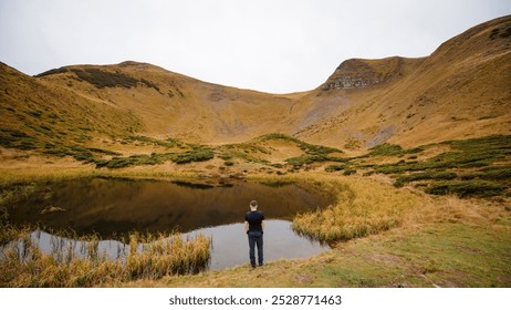 A person standing by a tranquil pond surrounded by rolling hills and grassy terrain. The landscape features a mix of green shrubs and golden grass under a cloudy sky, creating a serene atmosphere. - Powered by Shutterstock