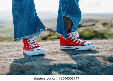 A person standing confidently on rocky terrain, wearing bright red sneakers and frayed denim jeans in the afternoon light - Powered by Shutterstock