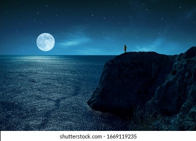 Person Is Standing In The Edge Of A Cliff Against The Sea And Full Moon, Under Stars And Moon Light.