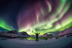 Person Standing In A Snowy Landscape With The Northern Lights In The Sky And A Lake In Front Of Him