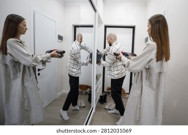 A person stands in front of a mirror using a hairdryer. Another person adjusts their outfit nearby. A cat sits on the floor in the background. The setting is a bright hallway with white walls.