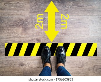 Person Stands In Line With Yellow And Black Stripes Keeping Safe Social 2m Distance, Arrow Due To Pandemic. Legs Of Girl In Jeans Waiting In Line At Checkout Counter In Supermarket Top View