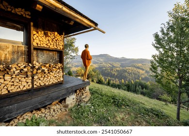 A person stands on the porch of a rustic cabin with stacked firewood, gazing thoughtfully at the serene mountain landscape during sunset - Powered by Shutterstock