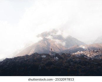 A person stands on a rocky terrain beneath a foggy mountain. - Powered by Shutterstock