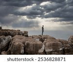 A person stands on weathered cliffs by the ocean under a moody sky. Sunlight pierces dark clouds, casting rays on the sea along Portugal