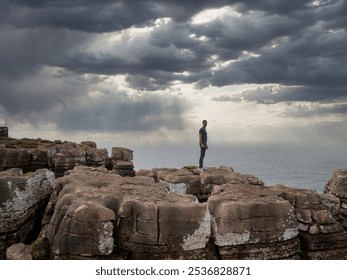 A person stands on weathered cliffs by the ocean under a moody sky. Sunlight pierces dark clouds, casting rays on the sea along Portugal's coast. - Powered by Shutterstock