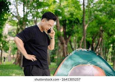 A person stands outdoors, engaged in a phone conversation while positioned next to a brightly colored camping tent amidst lush greenery and trees during daytime.
