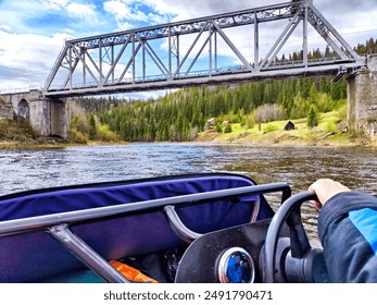 person steers a small motorboat under a steel bridge, navigating a calm river flowing through a lush forest - Powered by Shutterstock