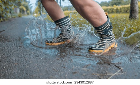 Person with striped rain boots jumping in a puddle of water, water splashing around on asphalt ground, grass and early autumn feeling around.