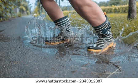 Person with striped rain boots jumping in a puddle of water, water splashing around on asphalt ground, grass and early autumn feeling around.