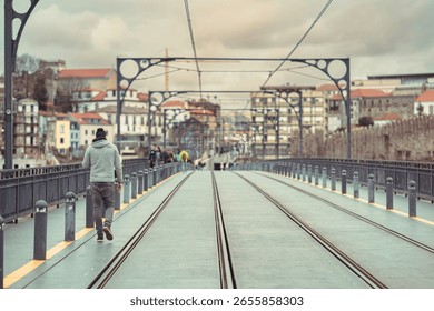 A person strolls across the upper deck of the Dom Luis I bridge, taking in views of Porto under a cloudy sky. The cityscape unfolds in the background, showcasing the urban charm