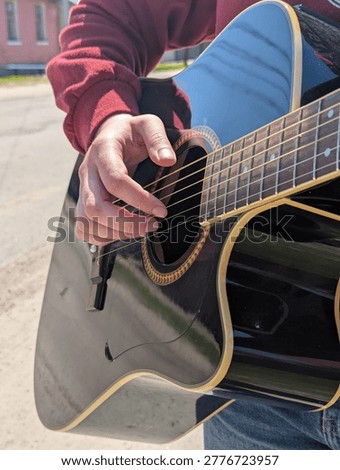 Person strums guitar strings while sitting outdoors on a sunny day in the neighborhood.