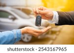 A person in a suit hand over a car key to another person with an open palm, symbolizing the transfer of ownership or rental of a vehicle. The background shows blurred cars, indicating a dealership.