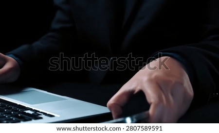A person in a suit using a smartphone and working on a laptop at a desk.