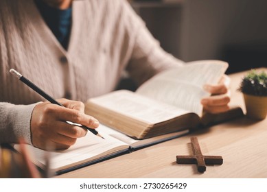 Person taking notes while reading an open Bible at a desk, pencil and journal beside a wooden cross, warm indoor light and soft focus, conveying Christian faith, devotion and scripture study at home.