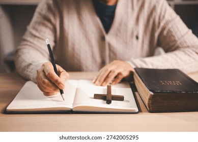 Person taking notes while reading an open Bible at a desk, pencil and journal beside a wooden cross, warm indoor light and soft focus, conveying Christian faith, devotion and scripture study at home.