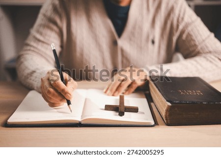 Person taking notes while reading an open Bible at a desk, pencil and journal beside a wooden cross, warm indoor light and soft focus, conveying Christian faith, devotion and scripture study at home.