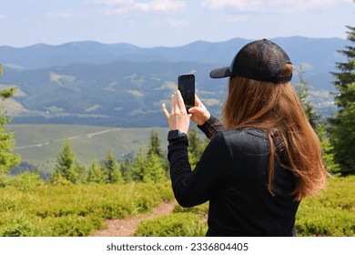 Person taking a photo with their phone of a scenic view. The person is standing on a grassy hill and is wearing a black baseball cap and a black long sleeve shirt. - Powered by Shutterstock