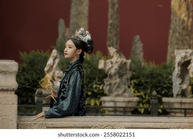 A person in traditional Chinese attire with a floral headpiece stands by a stone railing, holding a decorative fan in a landscaped setting. - Powered by Shutterstock