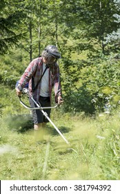 Person Trimming The Lawn With A Weed Eater Or Brush Cutter Wearing A Protective Helmet With Goggles To Shield His Eyes, Viewed Through Green Foliage.