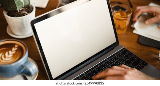 Person typing on a laptop with a blank screen, next to a coffee cup and a glass of juice on a wooden table at a cafe. Laptop, coffee, and typing scene. Blank laptop screen at cafe. - Powered by Shutterstock