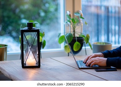 Person Typing On Laptop Keyboard At Kitchen Table. Working From Home During Winter Season With Candle Lantern On Wooden Table To Create Cozy Atmosphere Indoors.