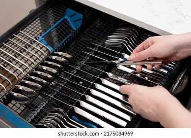 Person unloading open dishwasher machine with clean stainless cutlery set: knives, spoons and forks. Inside dishwashing machine with cutlery tray