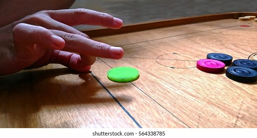 A Person Uses His Fingers To Flick A Green Striker Seed To Hit And Pocket A Playing Coin In A Carrom Game. The Red Queen Coin Must Be Pocketed And Subsequently Pocket A Carrom Man Of His Own Color. 